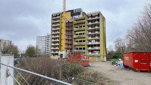 Ein mehrgeschossiges Wohnhaus mit grünem Anstrich ohne Fenster in einem verwahrlosten Zustand, umgeben von Baucontainern und Absperrungen.