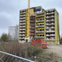 Ein mehrgeschossiges Wohnhaus mit grünem Anstrich ohne Fenster in einem verwahrlosten Zustand, umgeben von Baucontainern und Absperrungen.