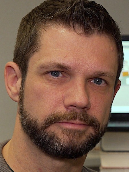 Frank Schulz, ein braunhaariger Mann mit Vollbart sitzt vor einem Bildschirm und lächelt freundlich.
