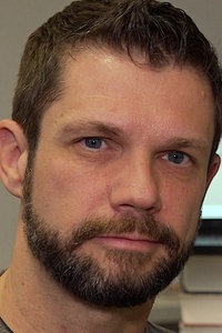 Frank Schulz, ein braunhaariger Mann mit Vollbart sitzt vor einem Bildschirm und lächelt freundlich.