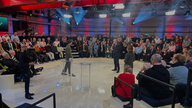 Bundeskanzler Friedrich Merz steht in der Mitte der "Arena"