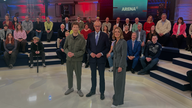 Bundeskanzler Freidrich Merz steht zwischen dem Moderations-Duo.