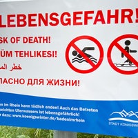 Ein Schild weist mit großen, roten Buchstaben auf verschiedenen Sprachen auf die Lebensgefahr beim Schwimmen im Rhein hin.