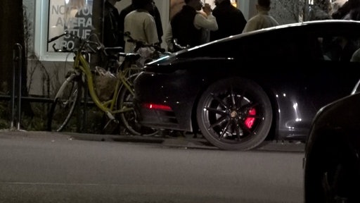 Menschen stehen vor einem Café in Köln Schlange, davor parkt ein augenscheinlicher Sportwagen