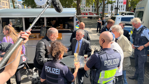 Herbert Reul steht mit zwei Polizisten und drei Männern um einen Tisch herum. Sie stehen vor einem Café-Wagen. Drumherum stehen weitere Polizisten und Polizeiwagen. Über den Tisch um den Herr Reul und die anderen stehen wird ein Mikrofon gehalten.