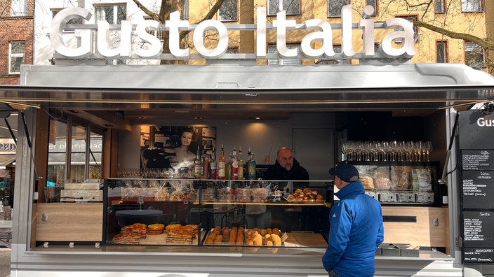 Bei einem Stand mit Italienischem Essen unterhalten sich der Standbesitzer und ein Kunde. 