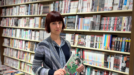 Eine Frau mit Pony-Frisur steht mit einem Buch in der Hand vor einem Bücherregal.