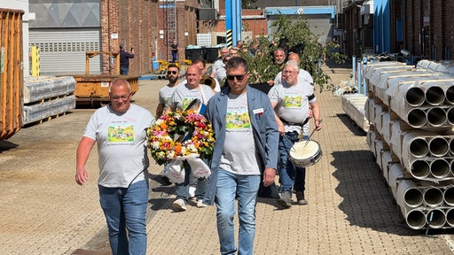 Die Mitarbeitenden des Unternehmens laufen in grauen T-Shirts hintereinander. Sie tragen Trommel, Blumenkranz und Maibaum.