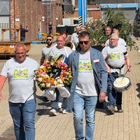 Die Mitarbeitenden des Unternehmens laufen in grauen T-Shirts hintereinander. Sie tragen Trommel, Blumenkranz und Maibaum.