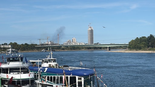 Bild vom Rhein. Im Hintergrund sieht man von einem Gebäude Rauschschwaden aufziehen.