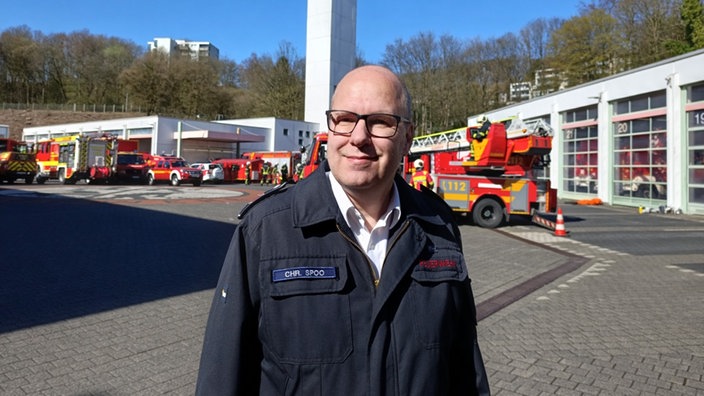 Ein Feuerwehrmann lächelt in die Kamera, dahinter die Wache und Einsatzfahrzeuge