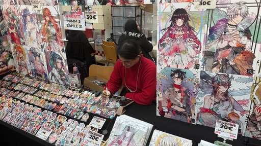 Ein Mann sitzt an einem Stand und zeichnet. Um ihn herum hängen viele Manga-Zeichnungen.