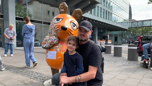Ein Vater kniet mit seiner Tochter im Arm vor der beschädigten Maus Figur