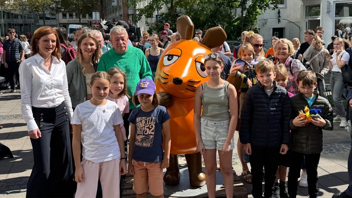 Einige Kinder und zwei Erwachsene stehen um eine orangene Mausfigur herum.
