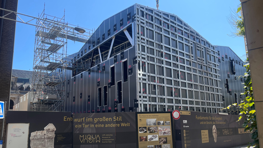 Eine unfertig Häuserfassade. Davor steht eine Absperrung mit der Aufschrift "MiQua - Entwurf im großen Stil, ein Tor in eine andere Welt"