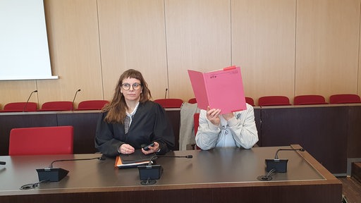 Der Angeklagte mit seiner Anwältin im Gericht. Er hält sich einen roten Ordner vor das Gesicht.