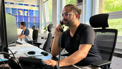 An einem Schreibtisch in einem Büro sitzt ein Mann mit Brille. In seiner rechten Hand hält er einen Telefonhörer.