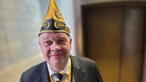 Ein lächelnder älterer Herr im Anzug mit goldener Narrenkappe und schwarz-gelber Krawatte