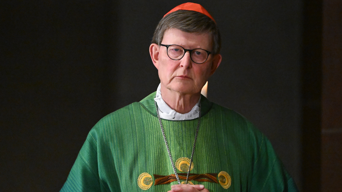 Ein Mann mit Brille und rotem Birett trägt ein grünes Gewand und blickt in die Kamera.