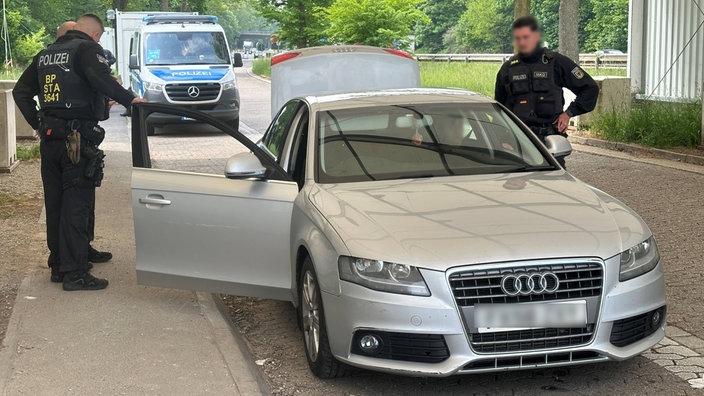 Auf dem Bild ist ein grauer Wagen und mehrere Polizisten. Im Hintergrund ist ein Polizeibulli.