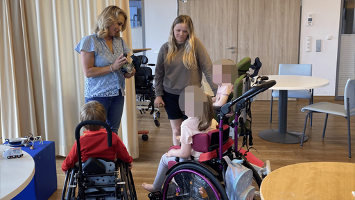 Zwei Frauen stehen in einem Raum. Vor ihnen sind drei Kinder in Rollstühlen zu sehen.