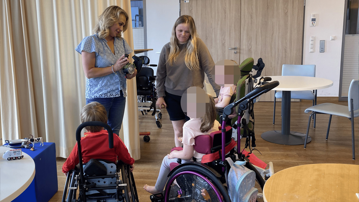 Zwei Frauen stehen in einem Raum. Vor ihnen sind drei Kinder in Rollstühlen zu sehen.