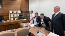 Vier Menschen in einem Gerichtssaal. Zwei davon sind unkenntlich gemacht. Die anderen beiden, ein Mann und eine Frau tragen Anwaltsroben.