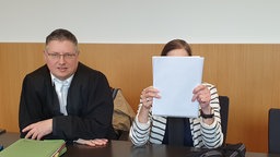 Zwei Personen sind in einem Gerichtssaal, eine Frau hält sich eine Mappe vor ihr Gesicht