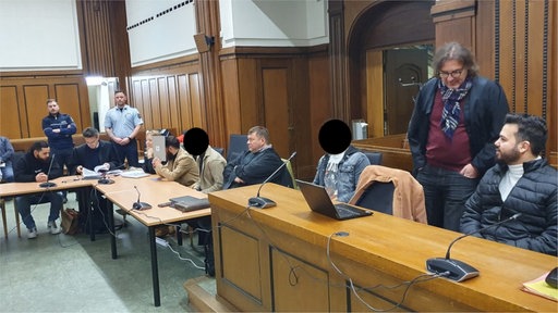 Die sechs Angeklagten sitzen mit ihren Verteidigern im Gerichtssaal. 