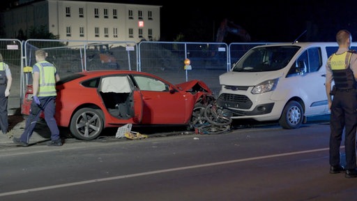Roter Sportwagen mit verbeulter Motorhaube. Zerquetschtes Fahrrad davor und direkt am Fahrrad ein kleiner weißer Transporter