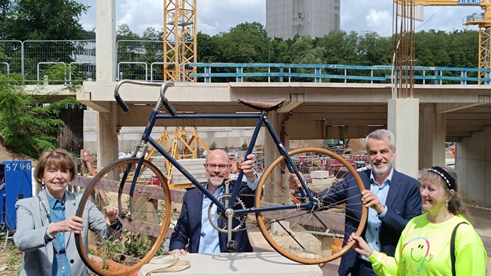 Vier Personen stehen auf einer Baustelle und halten gemeinsam ein altes, historisches Fahrrad in die Höhe. Im Hintergrund sind Betonpfeiler, Baugerüste und Kräne zu sehen