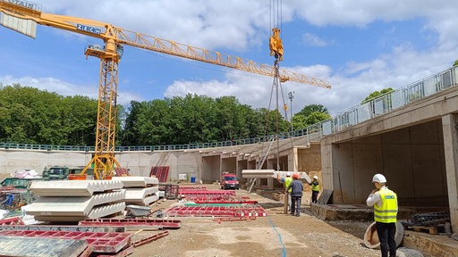 Auf einer Baustelle mit gelbem Baukran wird ein großes Betonteil mit Seilen angehoben. Mehrere Bauarbeiter in Sicherheitskleidung und Helmen stehen in der Nähe und beobachten den Vorgang