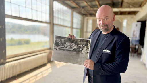 Mann steht im alten Hotel und zeigt ein Foto des Hotels von früher in die Kamera.