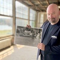 Mann steht im alten Hotel und zeigt ein Foto des Hotels von früher in die Kamera.