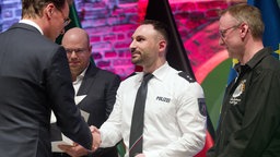 Hendrik Wüst (l.) überreicht Enver G. (m.) die Rettungsmedaille.