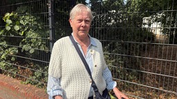 Karin Bramann steht mit einer Umhängetasche und einem weißen Oberteil vor einem Zaun auf einem Geheweg