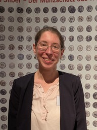 Vor einer Wand mit mehreren hundert Münzen drauf, steht eine Frau mit Brille. Sie schaut lächelnd in die Kamera.