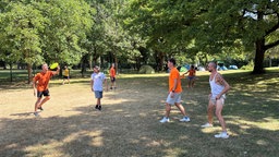 Vier Männer in Sportkleidung stehen auf einer Wiese im Schatten unter Bäumen und werfen sich eine gelbe Frisbee-Scheibe zu