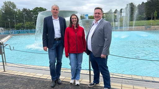 Vor einem Schwimmbecken stehen zwei Männer und eine Frau. Sie schauen lächelnd in die Kamera.