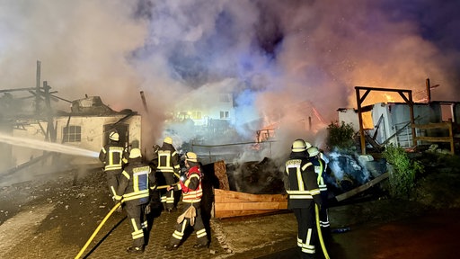 Feuerwehrleute stehen nachts mit Schläuchen ind er Hand vor abgebrannten Gebäuden eines landwirtschaftlichen Hofes.