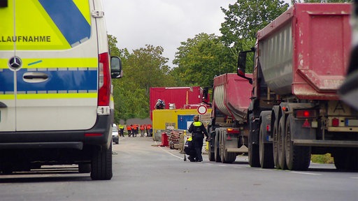 Rechts auf einer Fahrbahn stehen rote Lastwagen vor einer Baustelle, links daneben ein blau-weißer Polizeitransporter.