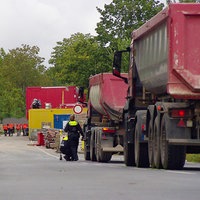 Rechts auf einer Fahrbahn stehen rote Lastwagen vor einer Baustelle, links daneben ein blau-weißer Polizeitransporter.