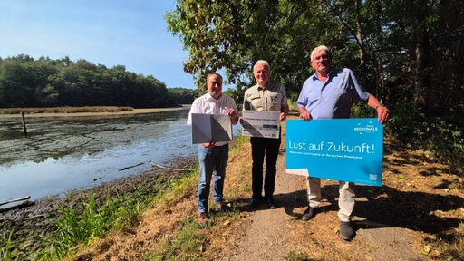 Drei Männer stehen neben einen Fluss und halten jeweils ein Schild hoch 