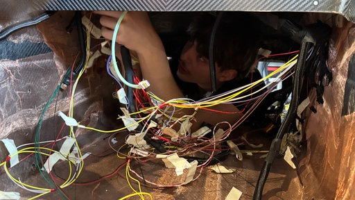 Kabel hängen im Innenraum eines Autos, ein Mechaniker guckt über Kopf durch ein Loch und befestigt sie. 