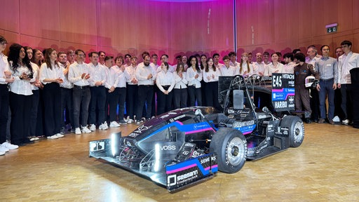 In weißen Hemden und Blusen und dunklen Hosen stehen Studierende um das selbst gebaute Rennauto. 