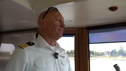An Mann in Kapitänsbekleidung und Sonnenbrille steht im Steuerhaus eines Schiffs