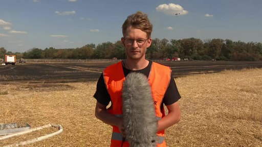 Ein Mann in orangener Warnweste und mit einem Mikrofon steht vor einem Waldgebiet.