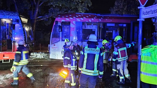 Zwei gekuppelte Straßenbahnen stehen im Hintergrund auf dem Gleis, davor stehen Feuerwehrleute, von Blaulicht beschienen