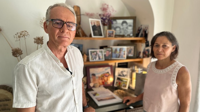 Ein Mann links und eine Frau rechts stehen vor einem Schreibtisch und Regalen voller Fotos, Kerzen und Erinnerungsstücken