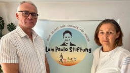 Links ein Mann, rechts eine Frau vor einem Plakat mit dem Logo der "Luis Paulo Stiftung" sowie dem Schriftzug "Mehr Schutz und Chancen für Kinder"
